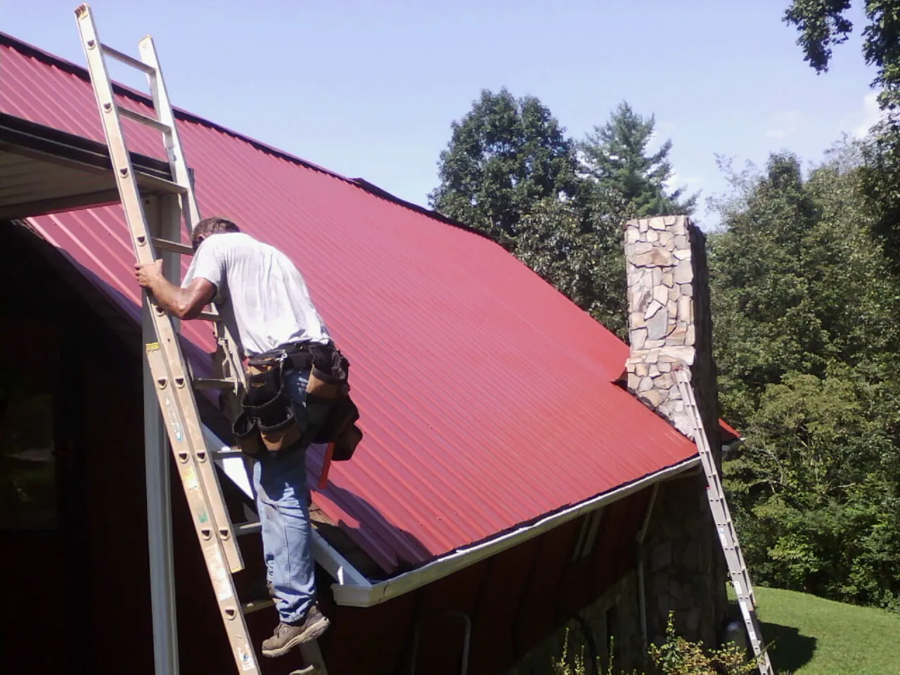 Roofer on a ladder beside a red metal roof and stone chimney — Soffit & Fascia Repair by Roofing Brothers in Barneveld